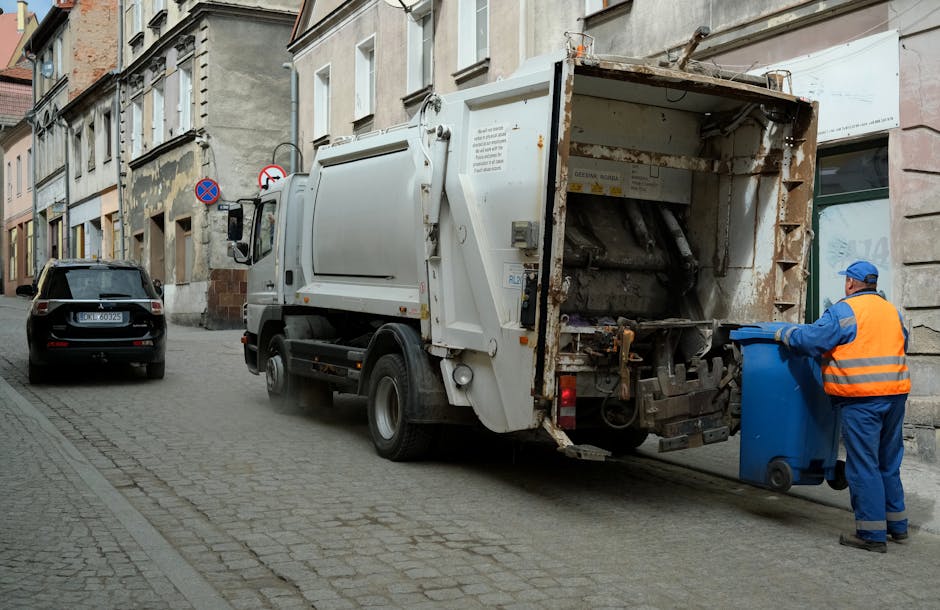 A street scene showing a waste collection vehicle operated by Man with Van Isle of Dogs parked next to a building on a cobblestone pavement, with an employee in a high-visibility vest and blue uniform standing by, emptying a blue wheelie bin into the truck's rear hopper during daylight hours on a cloudy day. The surrounding environment includes residential buildings with multiple stories, painted in light, neutral colors, and a few parked cars along the street. A large white waste truck is positioned with its rear hatch open, revealing rusted internal components used for compacting rubbish. The worker is focused on the task, holding the bin steady as it is being lifted and emptied into the truck. Nearby, there are cardboard boxes and other packing materials, suggesting a home relocation or moving process involving collection of bulky waste items, which aligns with house removals and furniture transport services provided by [COMPANY_NAME]. The scene indicates a typical scheduled waste pickup in an urban area, with professional staff handling waste removal efficiently as part of a broader move or clearing service.