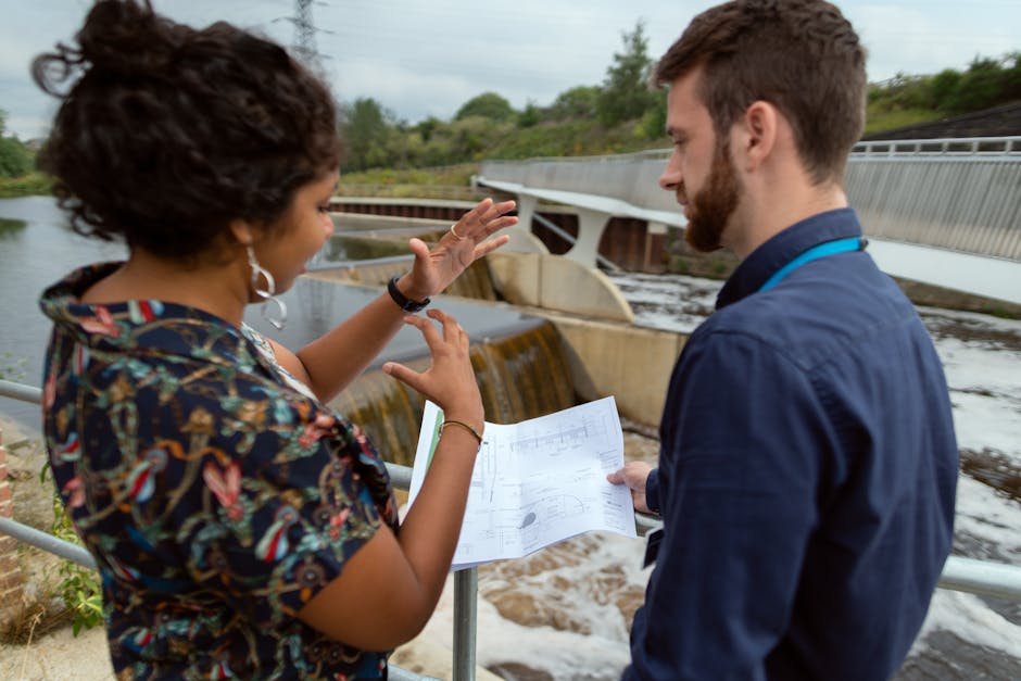 A woman with curly hair, wearing a colorful patterned blouse and earrings, is gesturing with her hands while speaking to a man with short brown hair and a beard, dressed in a navy blue shirt with a blue lanyard around his neck. They are standing on a paved area beside a waterway with a small waterfall or spillway in the background. A bridge spans across the water, with green trees and shrubs visible beyond. The woman holds a sheet of paper or blueprint, and the man is holding a similar document, both appearing engaged in a discussion about a project. The scene is outdoors during daytime with natural lighting, and the setting suggests a site visit or planning discussion related to a home or property development, akin to those involved in house removals or relocation planning that Man with Van Isle of Dogs might coordinate.