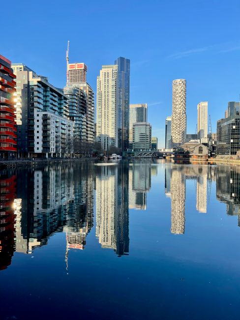 A clear daytime view of a modern city skyline reflecting on a calm river. The skyline features high-rise office buildings and residential towers with glass facades, some with unique architectural desi