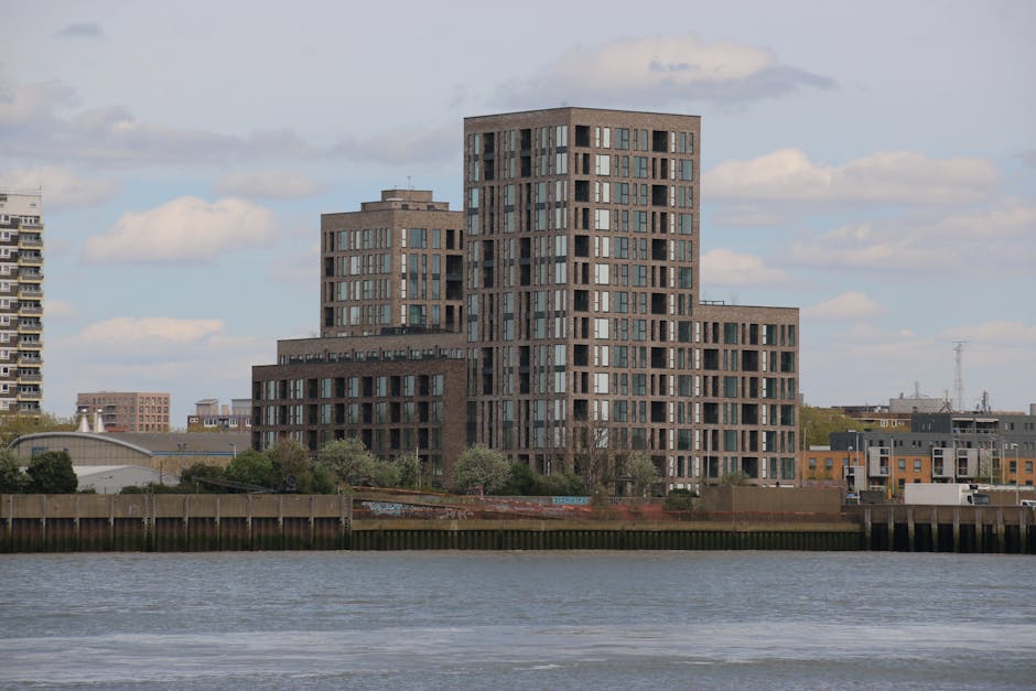 A view of a modern urban riverside scene showing high-rise office and apartment buildings with glass facades and geometric designs in the background, along with mid-rise structures and colorful reside
