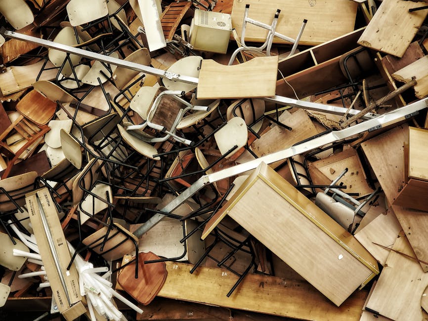A large pile of discarded furniture and various household items, including wooden desks, tables, chairs with metal and wooden frames, and plastic or fabric-covered chairs, all scattered and stacked haphazardly inside a property, likely an overloaded or hoarded flat on the Isle of Dogs. Some items are overturned or damaged, with chairs piled on top of each other and desks leaning against other objects. The scene includes cardboard boxes and packaging materials, with some furniture parts partially disassembled. The clutter is situated on a floor covered in dust and debris, with no clear pathways visible. This image reflects a home environment in need of clearance, illustrating the complexity of moving large quantities of furnishings and personal belongings. The scene conveys the importance of professional removals services like those provided by Man with Van Isle of Dogs to assist in loading, transporting, and clearing such overloaded spaces during a relocation process.