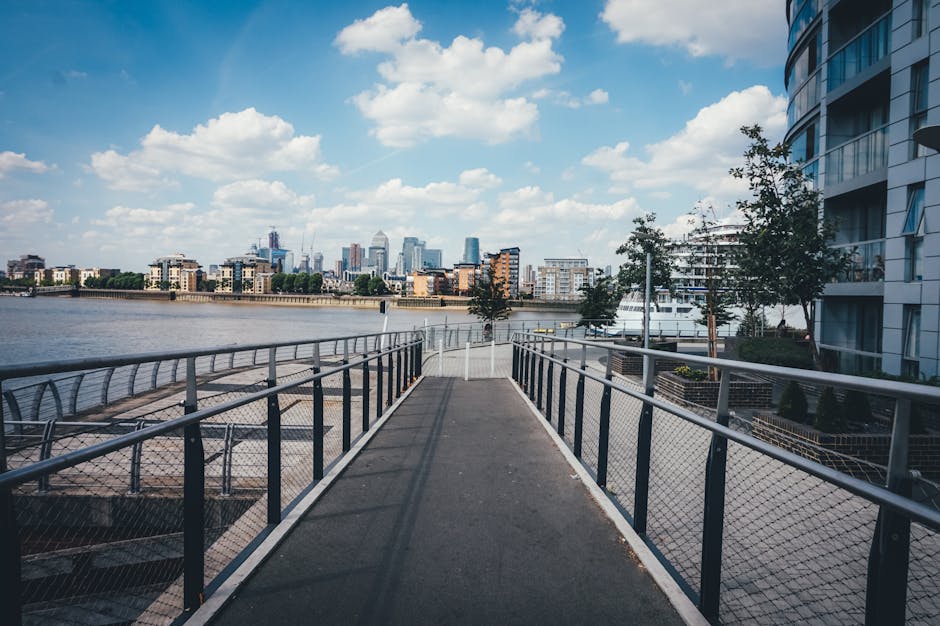 A straight, narrow concrete pathway with metal railings on both sides, situated on a riverside walkway in an urban area. The pathway is elevated over a lower promenade that features outdoor seating and a chain-link fence, with a city skyline featuring modern high-rise buildings visible across the river. To the right along the pathway, there is a tall, curved modern residential building with balconies, and small trees planted along the edge. The sky above is partly cloudy with patches of blue, and a boat is docked on the river on the right side of the image. The scene suggests a peaceful setting suitable for house removals or moving services within a city environment, with the image capturing a typical urban waterfront area during daytime.