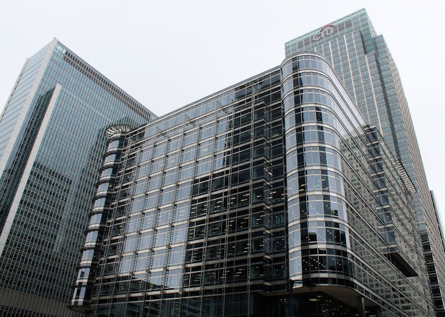 A cluster of modern high-rise buildings with glass facades and steel structures, located in an urban cityscape near Canary Wharf on the Isle of Dogs. The tallest building in the center has a curved, c
