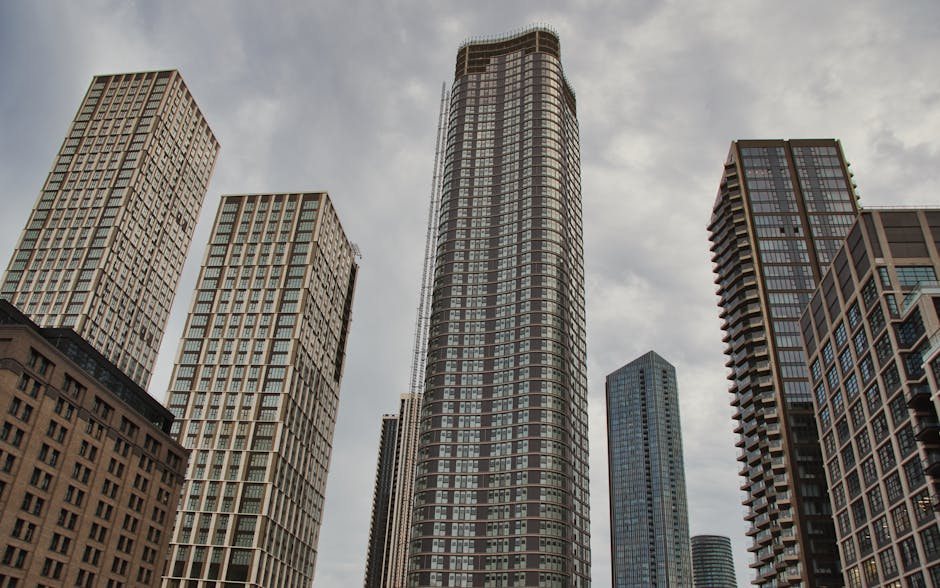 A cluster of modern high-rise buildings in an urban setting, viewed from ground level against a cloudy sky. The central skyscraper has a rounded, cylindrical shape with a facade consisting of horizontal bands of glass windows and dark-colored panels. Flanking it are other tall buildings with rectangular designs, featuring grid-like window patterns and varying exterior materials, including glass and concrete. The surrounding structures include one with a stepped roofline and another with a series of balconies on its facade. The scene depicts a cityscape typical of a financial district or commercial area, with no visible vehicles or people, focusing solely on the architecture and verticality of the buildings. This image may relate to home or business relocation services, such as those offered by Man with Van Isle of Dogs, emphasizing transportation and moving logistics within urban environments.