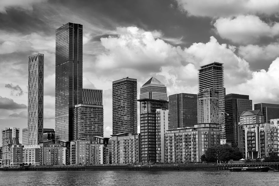 A black-and-white photograph of a modern city skyline showing high-rise office buildings and residential blocks along a waterfront, with some buildings featuring large glass facades and unique structural designs. The scene captures the urban environment during daylight, with a partly cloudy sky overhead. The image depicts a dense cluster of tall buildings behind lower residential structures, typical of a central business district within the Isle of Dogs area. This image aligns with house removals and relocating services performed by Man with Van Isle of Dogs, emphasizing urban furniture transport, loading processes, and the logistical challenges of moving in a cityscape.