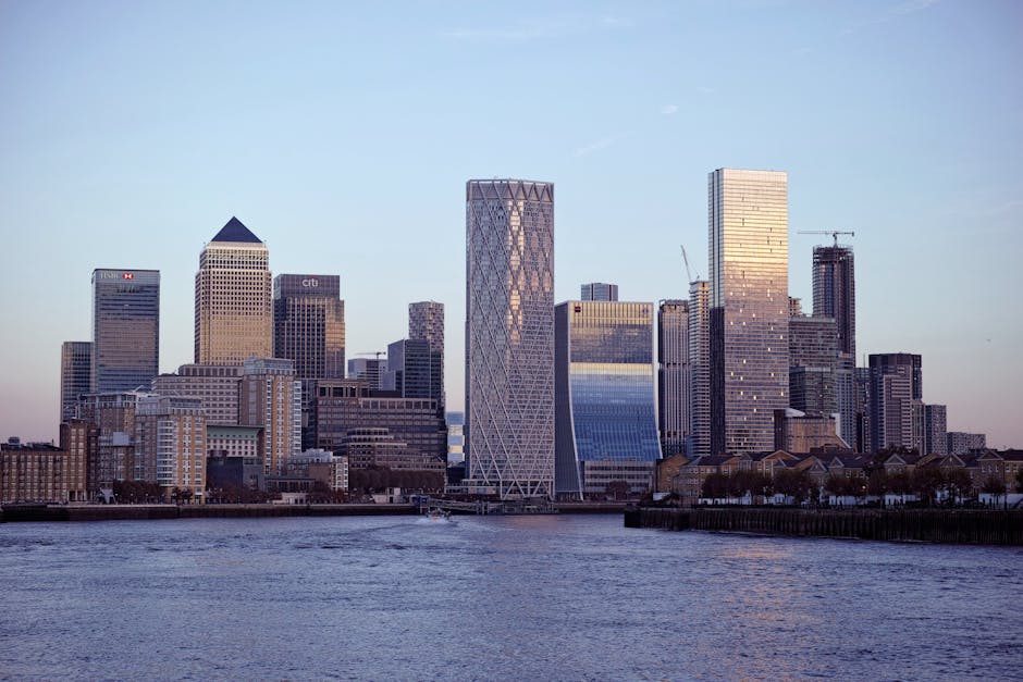 A cluster of modern high-rise buildings in an urban setting, viewed from ground level against a cloudy sky. The central skyscraper has a rounded, cylindrical shape with a facade consisting of horizontal bands of glass windows and dark-colored panels. Flanking it are other tall buildings with rectangular designs, featuring grid-like window patterns and varying exterior materials, including glass and concrete. The surrounding structures include one with a stepped roofline and another with a series of balconies on its facade. The scene depicts a cityscape typical of a financial district or commercial area, with no visible vehicles or people, focusing solely on the architecture and verticality of the buildings. This image may relate to home or business relocation services, such as those offered by Man with Van Isle of Dogs, emphasizing transportation and moving logistics within urban environments.