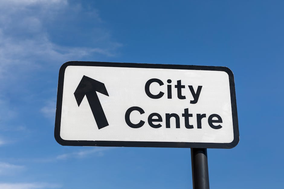 A rectangular road sign mounted on a black pole against a clear blue sky, with bold black text reading 'City Centre' and an upward-pointing arrow symbol indicating the direction. The sign is located outdoors, likely on a street or near a transport route, providing directional guidance. The image is relevant to house removals and packing and moving services offered by Man with Van Isle of Dogs, especially when navigating urban areas such as Westferry Road in E14, Isle of Dogs, during a home relocation or furniture transport process.
