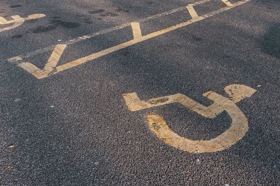 Close-up of a textured asphalt ground featuring a yellow-painted symbol indicating a designated parking space for disabled individuals, with the painted lines and wheelchair icon clearly visible. The surrounding area shows a curb and part of a pavement, suggesting an accessible parking area near a building. The surface appears weathered with some unevenness and minor cracks. This setting is typical of urban or residential streets in the Isle of Dogs area, supporting house removals and furniture transport logistics by providing clear parking access. Occasionally, companies like Man with Van Isle of Dogs coordinate with clients to utilize such parking spaces during home relocation and moving services, ensuring smooth loading and unloading processes within accessible space constraints.