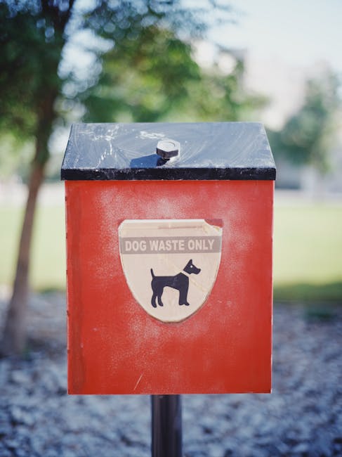 Close-up of a red and black outdoor dog waste disposal bin with a metallic lid, mounted on a black pole. The front of the bin features a label with a black silhouette of a dog and the text 'DOG WASTE ONLY' in grey and white. The background shows a blurred view of trees, grass, and a pathway, indicating a park or garden setting. The image captures the bin as part of an outdoor environment possibly near a residential area, relevant to home relocation or outdoor cleaning and waste management related to house removals and moving services provided by Man with Van Isle of Dogs.