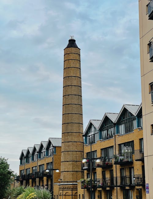 A tall, cylindrical chimney constructed from yellow brick with horizontal black bands elevates above modern residential buildings in Isle of Dogs. The nearby buildings are multi-storey, featuring yellow brick facades and rows of balconies with black railings, some decorated with potted plants and outdoor furniture. The scene is set outdoors during daytime with a partly cloudy sky overhead. In front of the buildings, there are street-level lights and some greenery. This image captures the urban environment involved in house removals and furniture transport, illustrating the typical setting for loading and unloading during home relocation or moving services. Man with Van Isle of Dogs may utilize such environments for loading furniture and boxes onto their vans as part of their packing and moving procedures, ensuring an efficient loading process within the residential area.