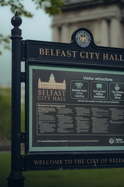 A close-up image of a black Belfast City Hall signage board, mounted on an iron post, displaying information about visitor attractions, guided tours, self-guided visits, and the Bobbin Coffee Shop. Behind the sign, lush green foliage and a statue are partially visible, indicating an outdoor setting. The sign features gold and white text and graphics, with a detailed illustration of Belfast City Hall's façade and the official Belfast City Council crest at the top. Although not directly related to house removals or moving services, this image illustrates the local context and environment of Isle of Dogs, relevant to regional relocation services provided by Man with Van Isle of Dogs.